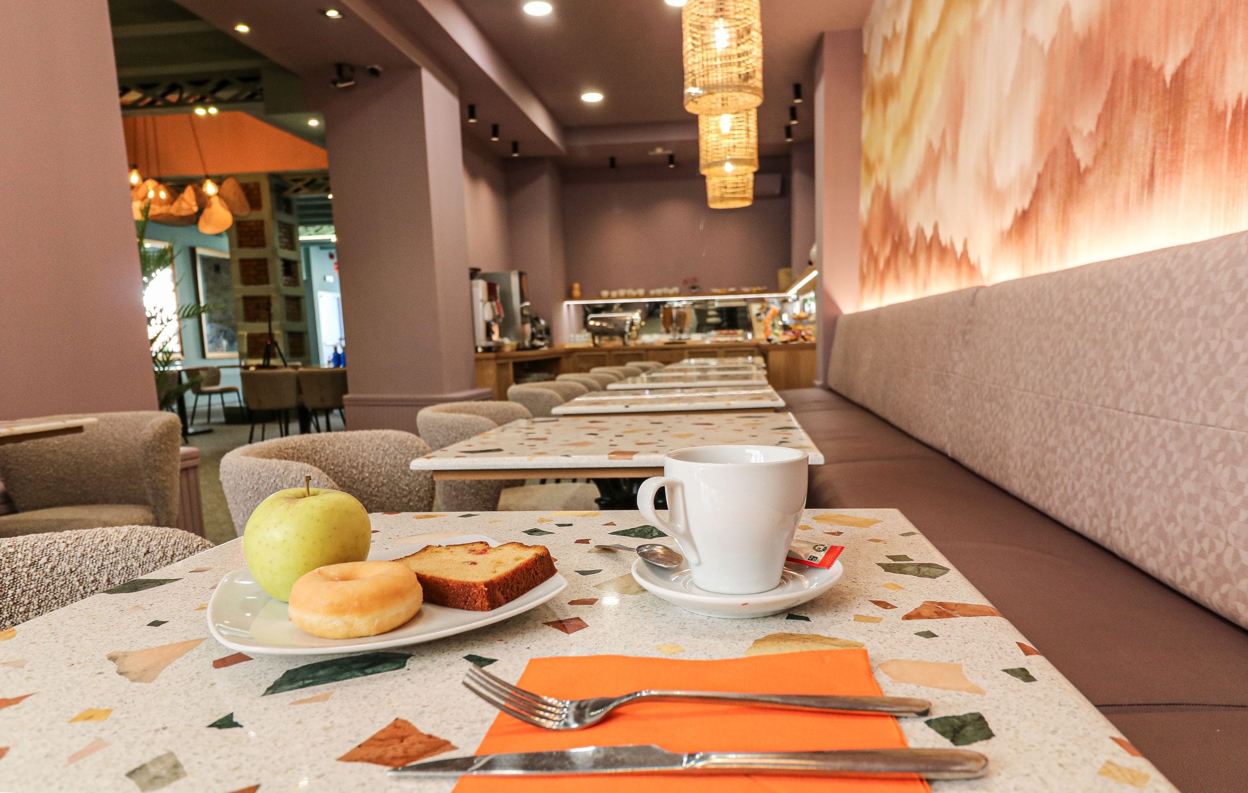 Zona de mesas en restaurante de diseño con sillas de tonos neutros, iluminación decorativa cálida y un mural pintado en tonos rosados y tierra.
