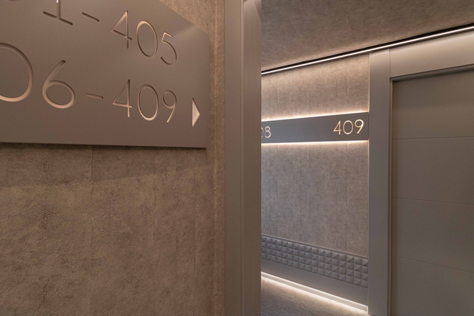 Pasillo de hotel contemporáneo con iluminación led indirecta superior y puertas de madera con indicadores numéricos luminosos.
