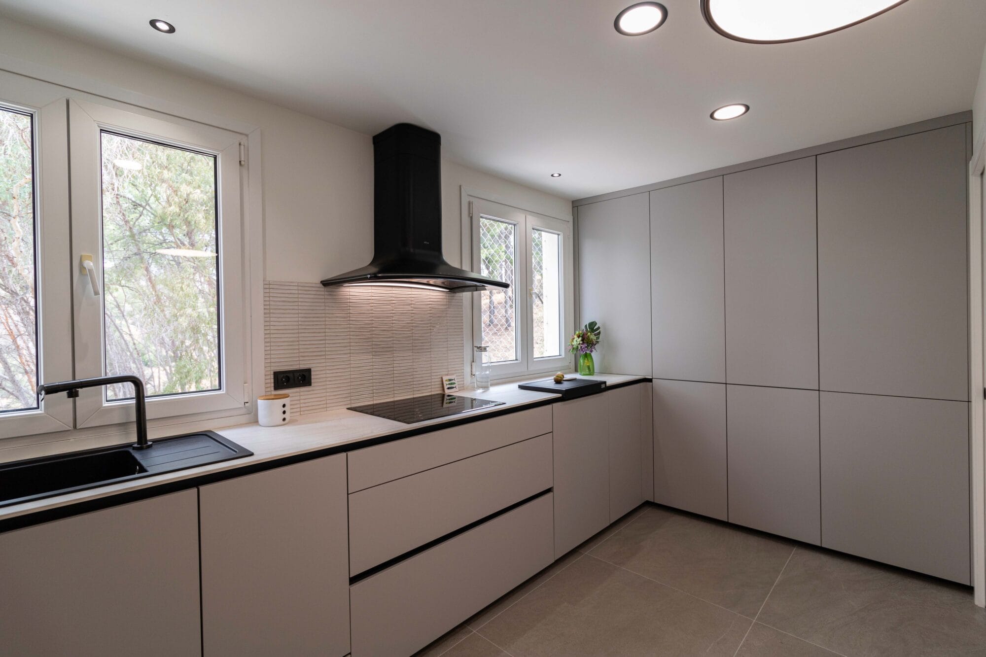 Diseño de cocina minimalista con muebles sin tiradores en tonos grises, ventanas de aluminio blanco y suelo porcelánico.