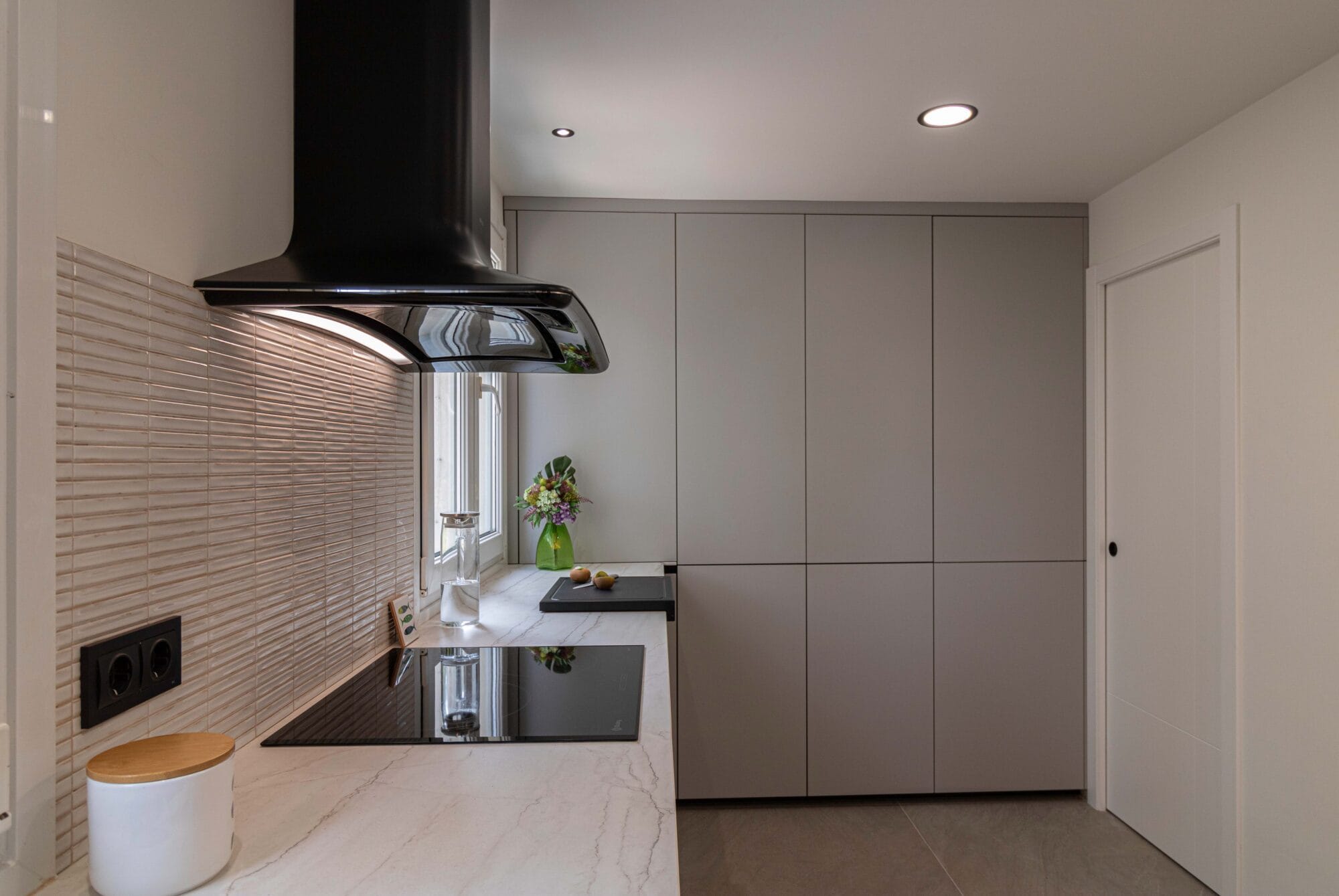 Reforma integral en Málaga de cocina moderna con encimera clara, placa de inducción y campana decorativa negra bajo iluminación LED integrada.