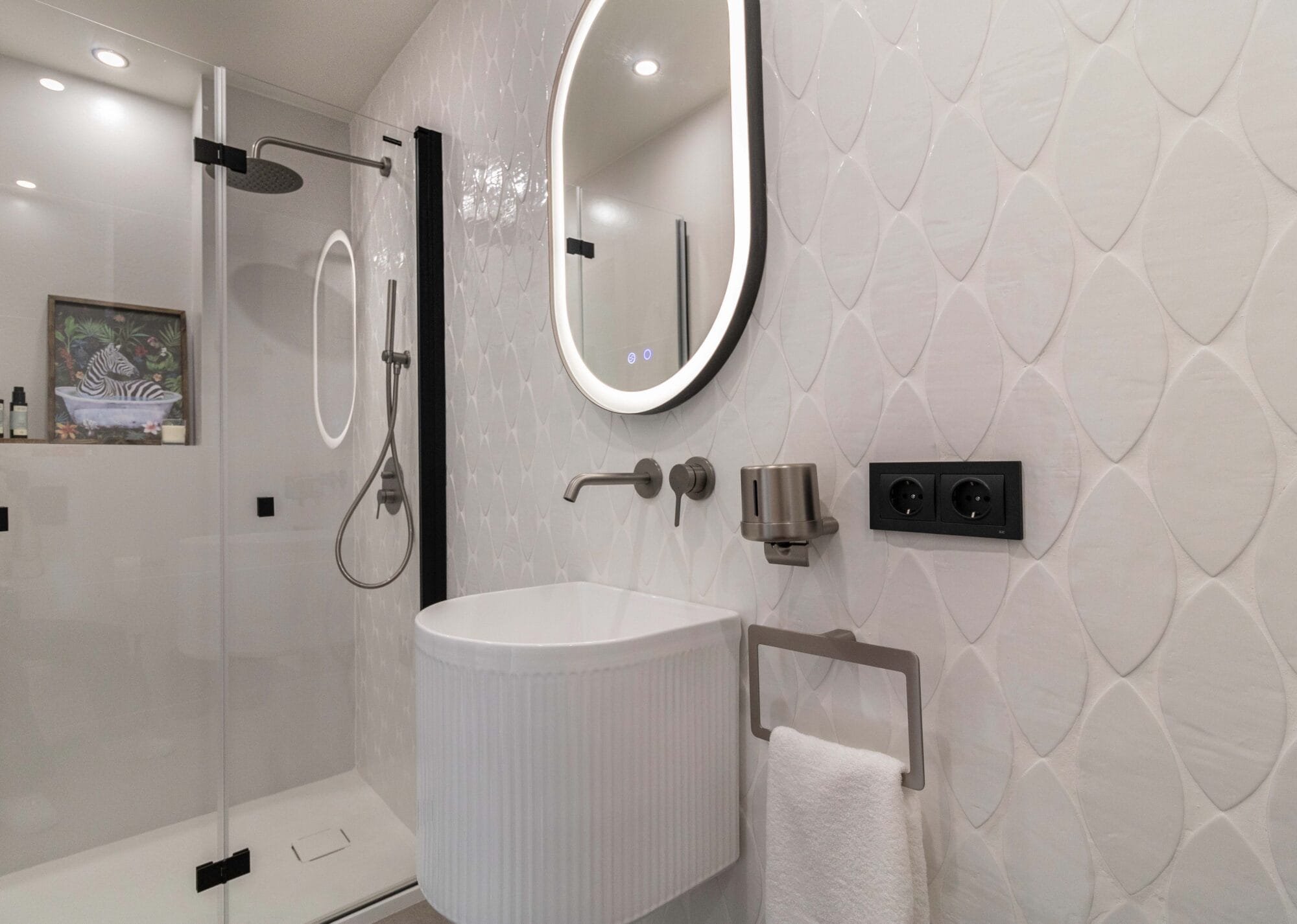Lavabo exento de diseño estriado en aseo moderno con espejo ovalado retroiluminado, grifería empotrada acero y azulejos con relieve en forma de hoja.