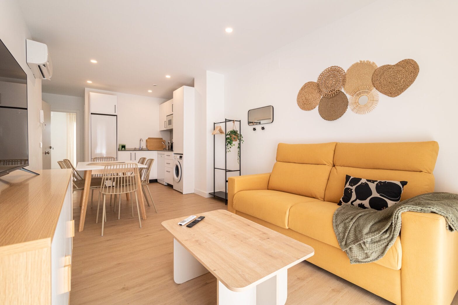 Diseño de salón comedor por interioristas en Málaga con un sofá amarillo mostaza, mesa de centro de madera, decoración de pared de mimbre y cocina al fondo.