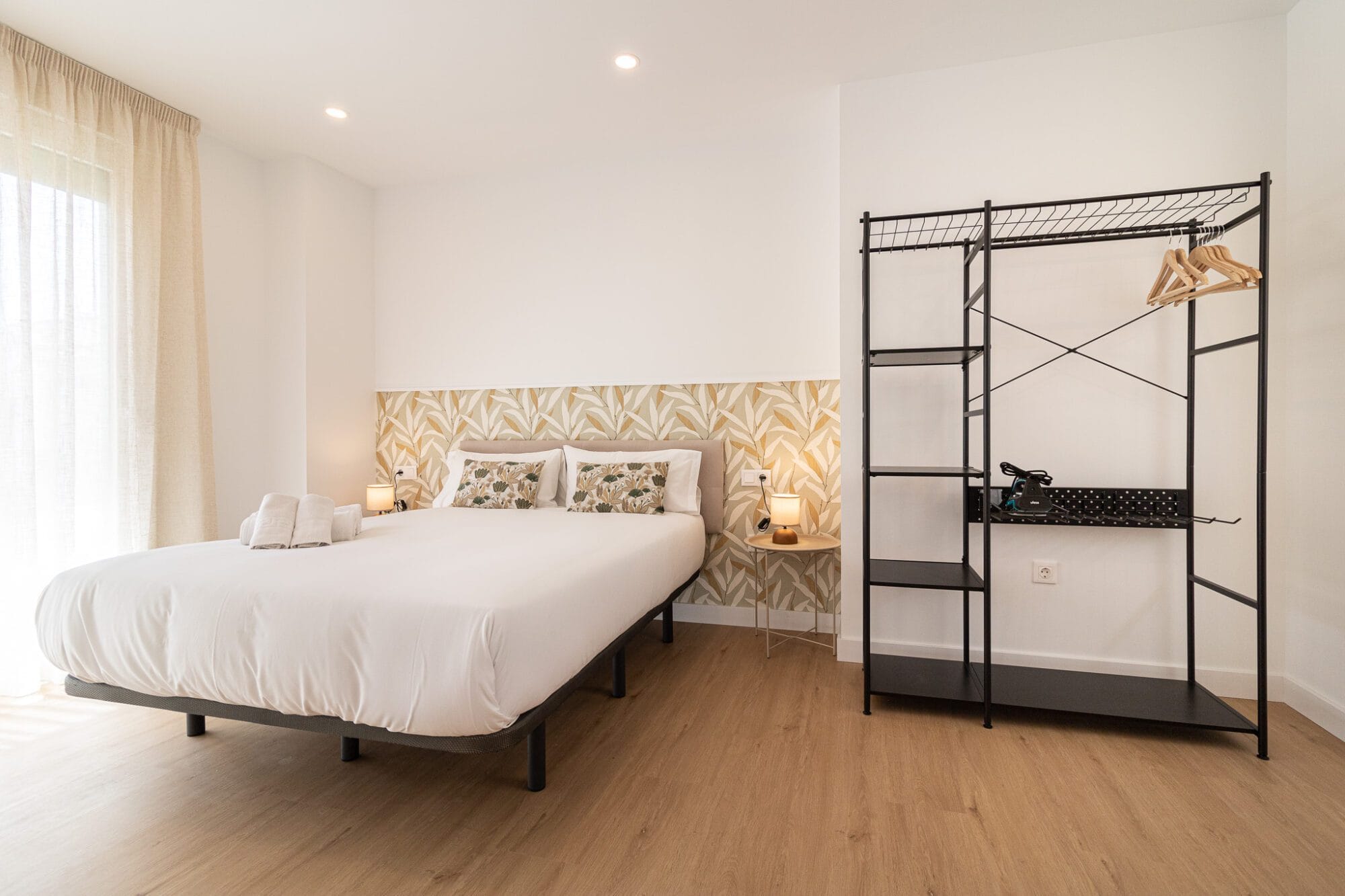 Diseño de interiores de un dormitorio moderno en Málaga con papel pintado de hojas doradas, cama de matrimonio y un perchero abierto de metal negro.