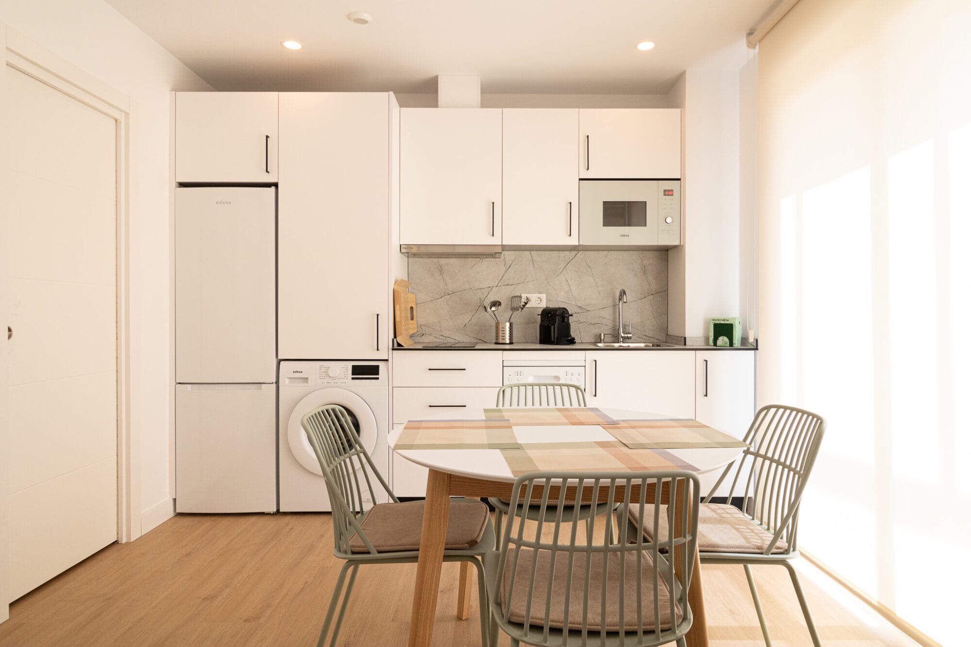 Proyecto de decoración de cocina lineal en color blanco con frente de efecto mármol gris y mesa de comedor de madera clara en un apartamento.